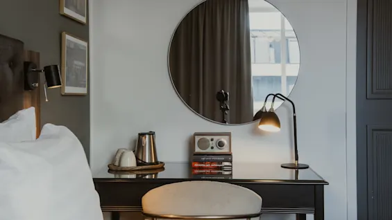 Tyylikäs hotellihuone, jossa on työpöytä, vedenkeitin, radio, kirjoja (JOHANNA MO, OM ANNA) ja lamppu. Pyöreä peili heijastaa ikkunan.