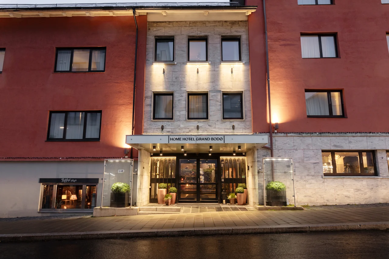 The illuminated entrance of a hotel building at dusk, with the sign HOME HOTEL GRAND BODO above the doors.