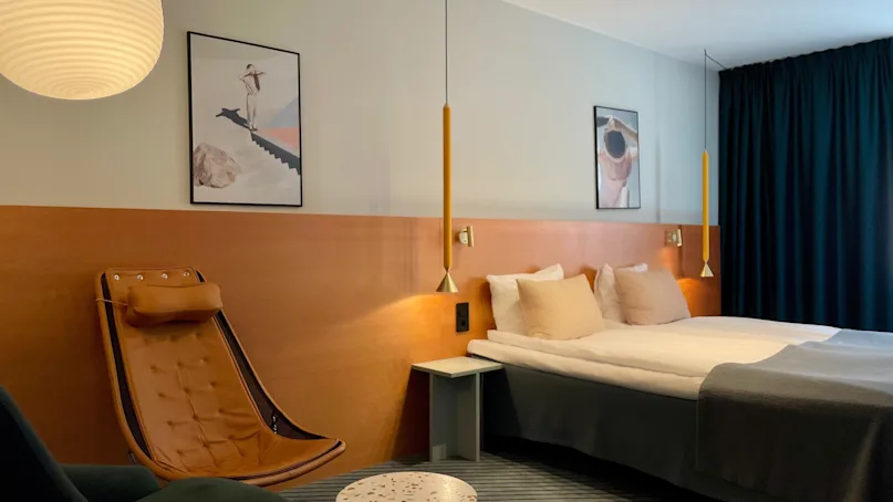 A modern hotel room with a double bed is framed by two hanging lamps. A leather chair is beside a small round table, and abstract art decorates the walls.