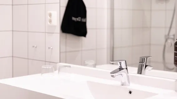 Et moderne badeværelse med en hvid flisevæg, en vask og en kromhane. En sort taske med teksten Hair Dryer hænger på væggen.