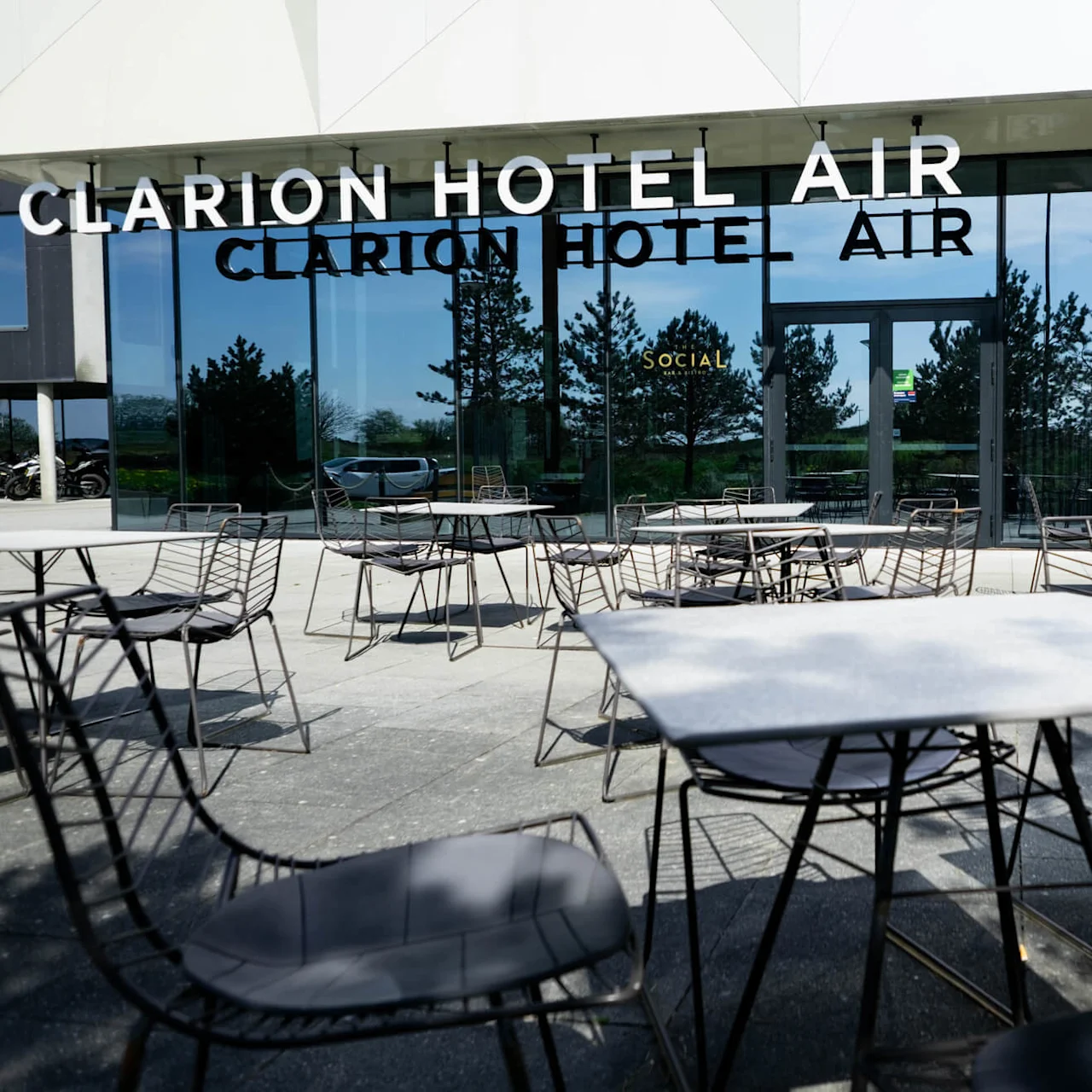Utendørsbord og stoler står plassert på en terrasse utenfor glassfasaden til Clarion Hotel Air. Trær reflekteres i vinduene. Synlig tekst: "CLARION HOTEL AIR" og "SOCIAL."