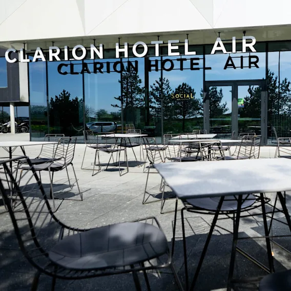 Utendørsbord og stoler står plassert på en terrasse utenfor glassfasaden til Clarion Hotel Air. Trær reflekteres i vinduene. Synlig tekst: "CLARION HOTEL AIR" og "SOCIAL."
