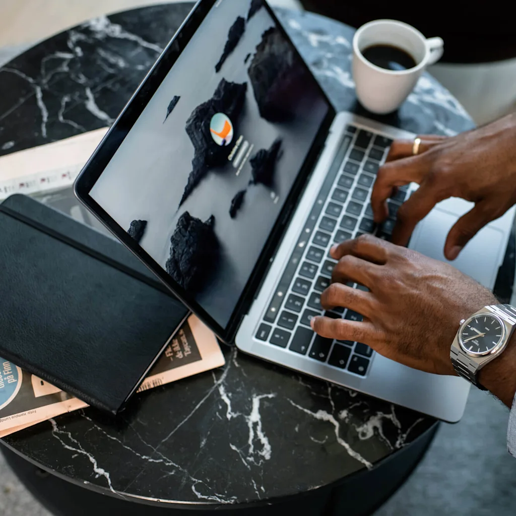En person skriver på en bærbar PC ved et marmorbord, med en notatbok, avis og kaffekopp. Skjermen viser en innloggingsside med kupert øylandskap.