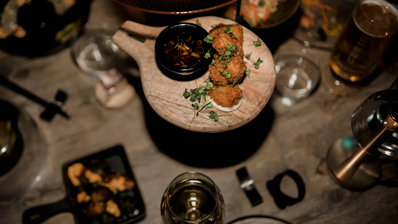 Brödpanerad friterad mat på en träbricka omgiven av olika rätter och ett glas öl, som avbildar en social måltidssituation.