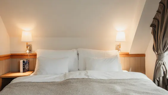 A cozy bedroom with a neatly made bed, white pillows, and a warm grey blanket, illuminated by soft wall lamps.