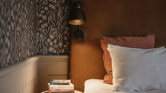 A cozy bedside scene with a warm lamp illuminating books on a nightstand next to a bed with pillows.