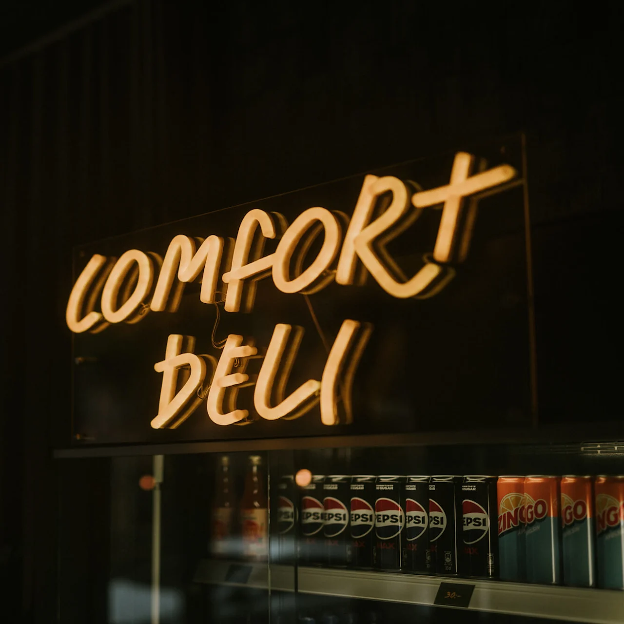 Et neonskilt med teksten "COMFORT DELI" over en drikkekøler i et dæmpet oplyst rum.