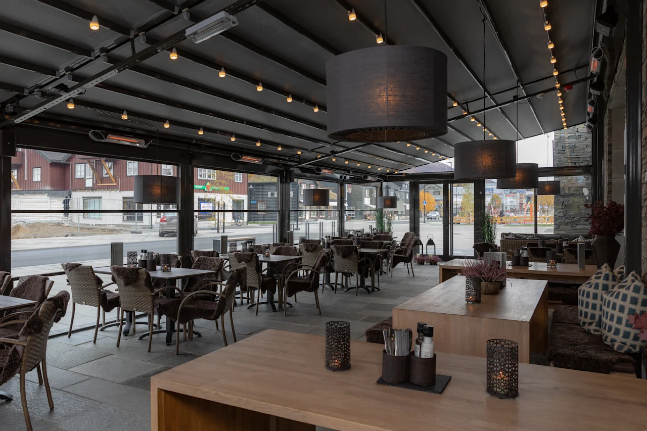 Interiør av en moderne restaurant med tomme flettede stoler og bord under et baldakintak. Hengende lys gir atmosfære, store vinduer gir utsikt over gaten utenfor.