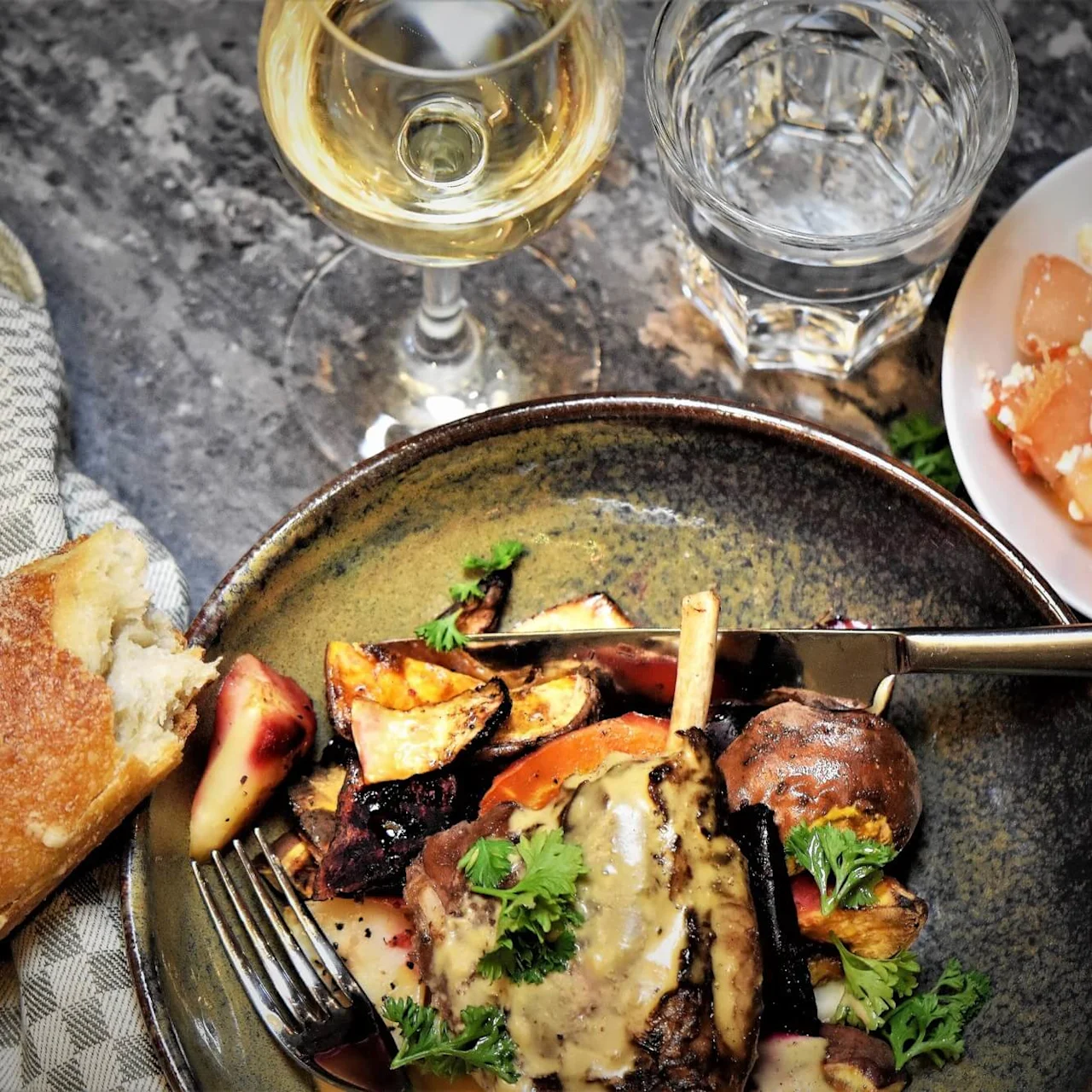 Et gourmetmåltid med stekte grønnsaker og kjøtt, glass med hvitvin, vann og brød på et flekkete bord.