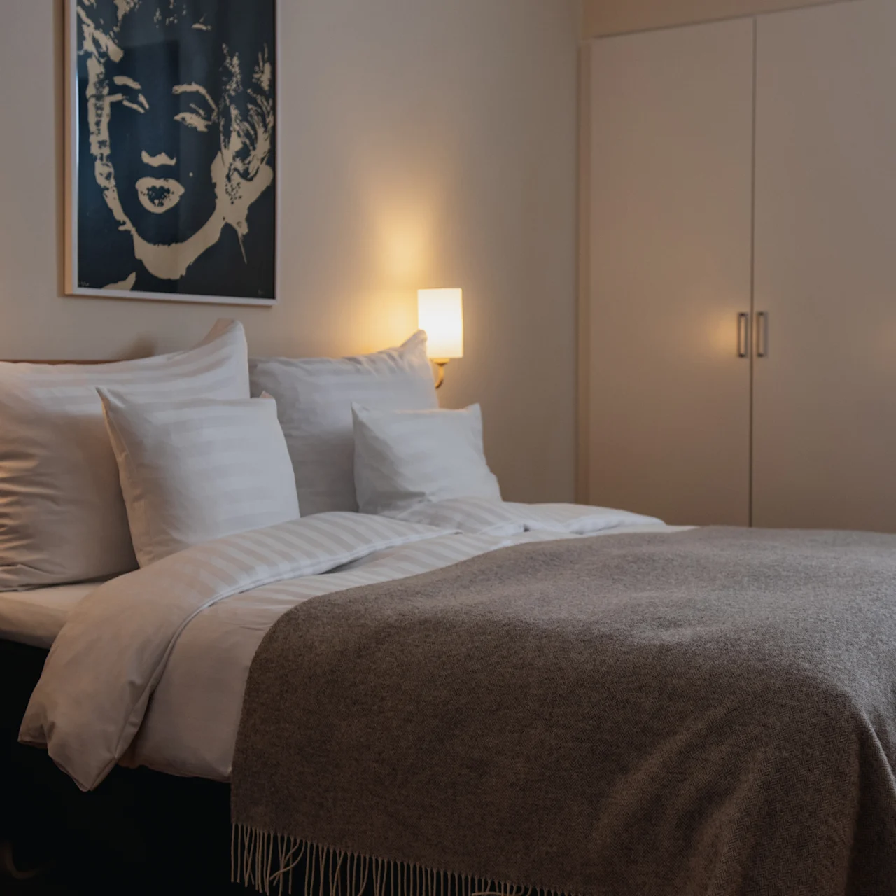 A neatly made bed with multiple pillows is illuminated by two wall-mounted lamps; a dark abstract portrait hangs above the bed in a cozy room with a wardrobe.