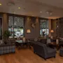 Koselig lounge med rutete lenestoler og sofaer arrangert rundt trebord, opplyst av varme gulvlamper. Veggene har firkantede dekorative aksenter og hjortegevir. Store vinduer viser en utendørs sitteplass.