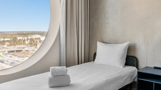 Et moderne hotelværelse med en enkeltseng, pænt foldede håndklæder og et markant cirkulært vindue med udsigt over en pulserende by.