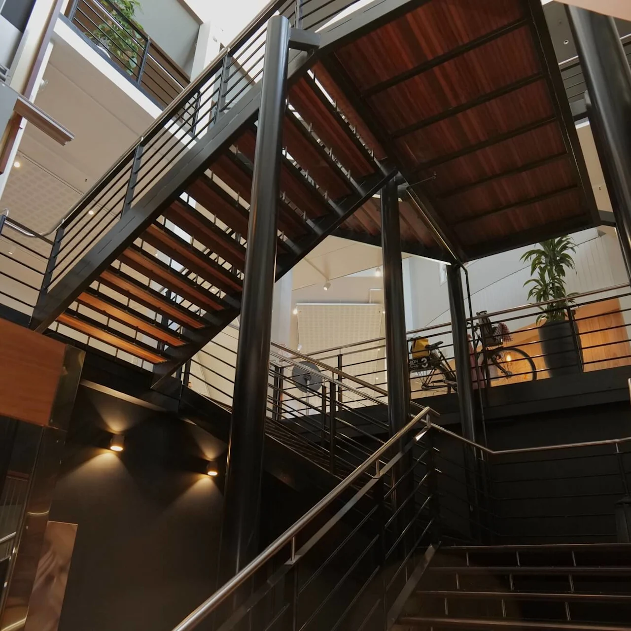 Danish: Trappe stiger omgivet af metalgelænder i et moderne bygningatrium, flankeret af plante på øverste niveau, med elevatorens etagenummer "2" synligt.