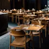 Bord som är elegant dukade med glas och servetter, upplysta av små lampor, är arrangerade i en dämpad, exklusiv restaurang. Trästolar omger varje bord, vilket förstärker den mysiga stämningen.