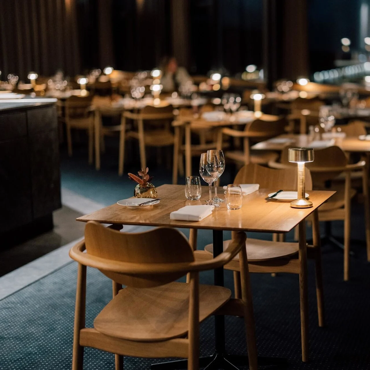Borde er elegant dækket op med glas og servietter, oplyst af små lamper, arrangeret i en svagt oplyst, eksklusiv restaurant. Træstole omgiver hvert bord, og forbedrer den hyggelige stemning.