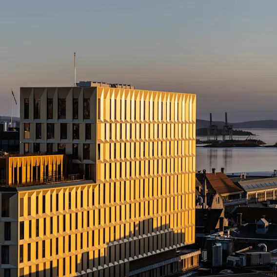 Golden-hued building casts shadows, surrounded by other buildings in urban area, with calm water and industrial cranes visible in the distant background.