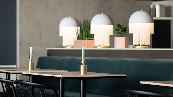 Bord med gullbase lamper er plassert i en moderne kafé. Over henger hyller med planter i rosa bokser mot en betongvegg, noe som tilfører grønt til den stilige settingen.