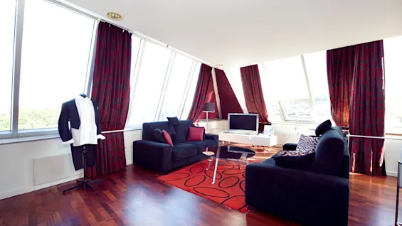 A modern living room with large windows, a red-patterned area rug, dark sofas, and a mannequin with a suit.
