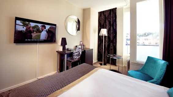 A hotel room with a bed, mounted TV displaying a show with subtitles "Jo, mye, men ikke noe spesielt.", and a window view of a waterfront.