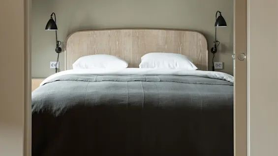 A bed with a wooden headboard and gray bedding, flanked by two black wall-mounted lamps, is centered in a small, minimalist room.