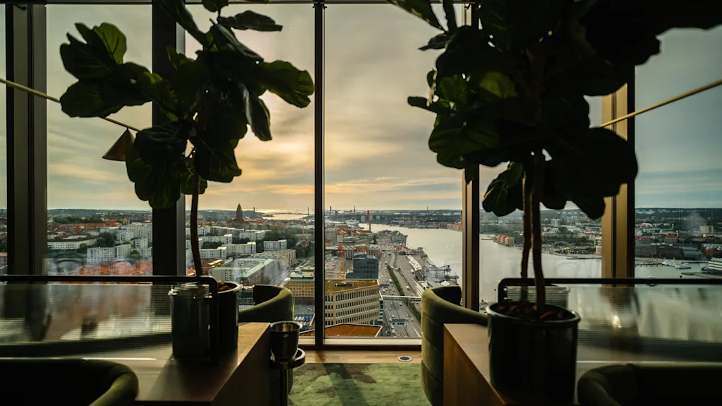 To potteplanter står på et bord ved gulv-til-tak-vinduer med utsikt over en bylandskap med vann, bygninger og en skyet himmel ved solnedgang.