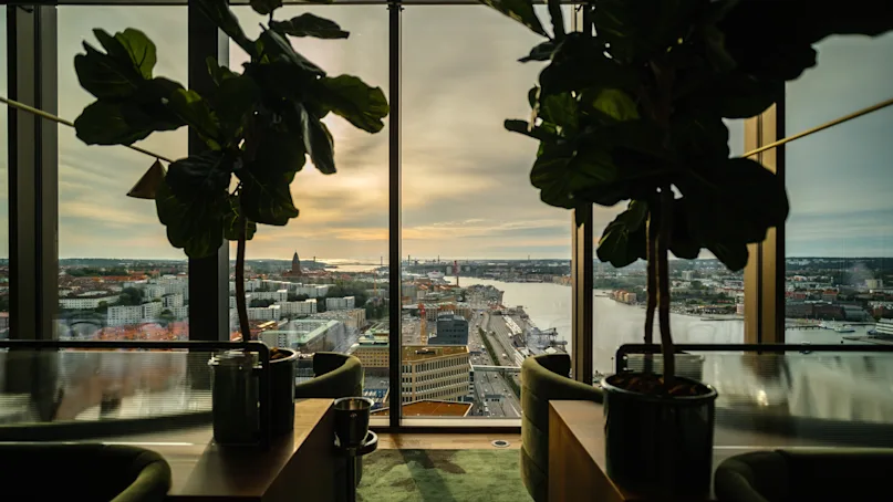 Two potted plants sit on a table by floor-to-ceiling windows, overlooking a cityscape with water, buildings, and a cloudy sky at sunset.