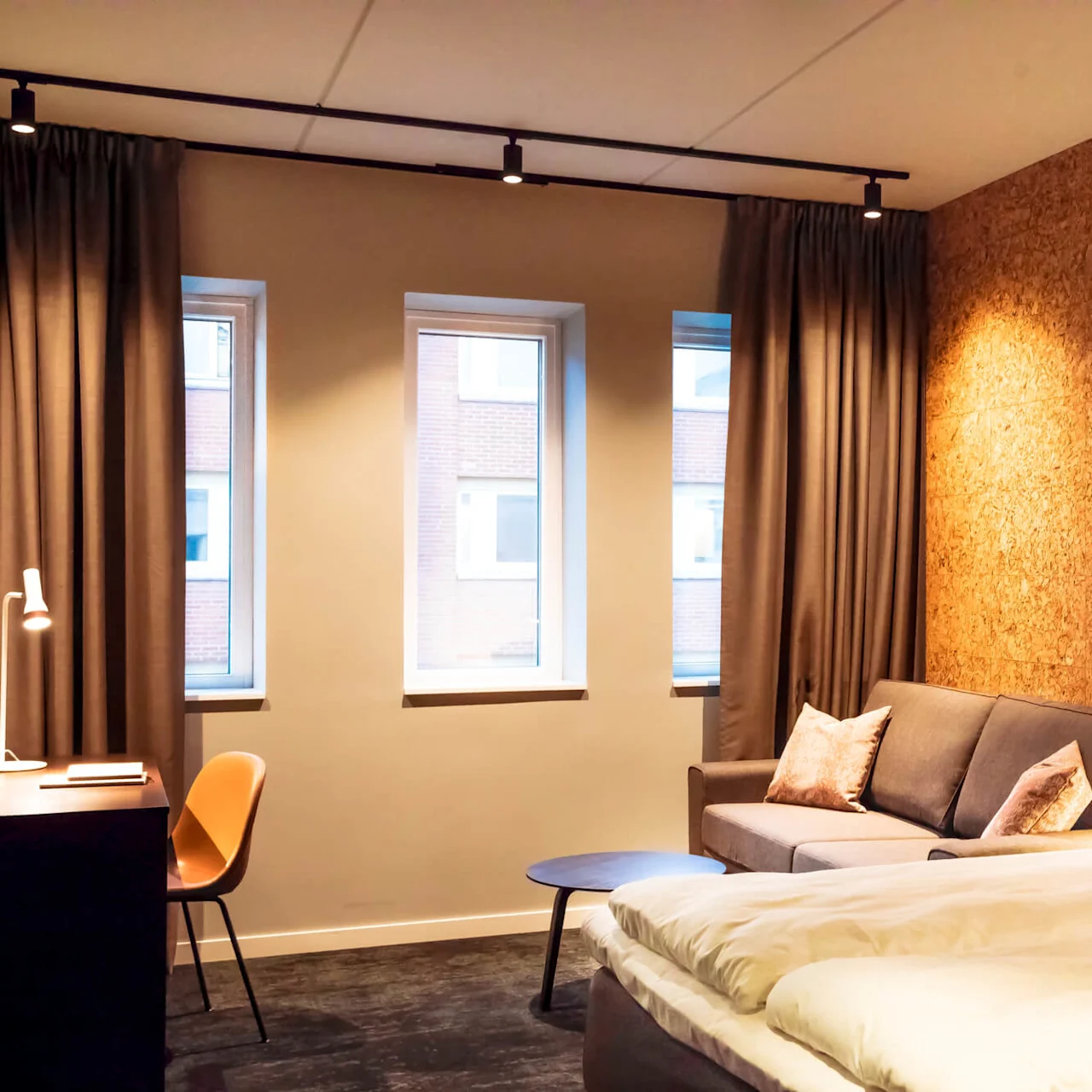 Bed with white linens sits against a cork wall, next to a gray couch in a warmly lit room. Three small windows reveal a brick building outside.
