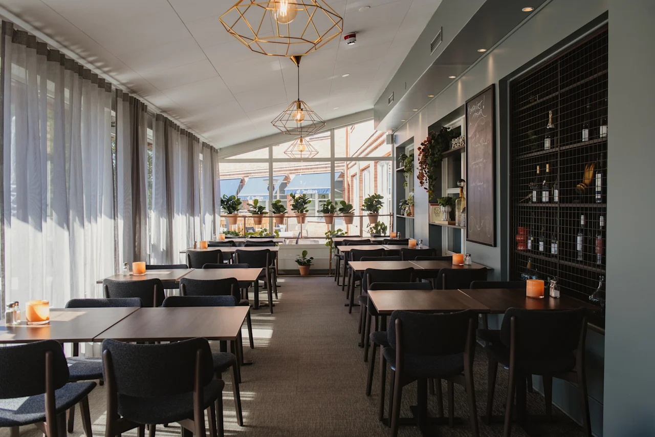 En välbelyst restaurangmatsal med flera bord redo för gäster, med stora fönster och krukväxter.