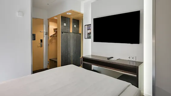 En flat-TV är monterad på en vägg ovanför ett skrivbord i ett modernt hotellrum. Nära finns en garderob av trä och en stängd dörr i minimalistisk design.
