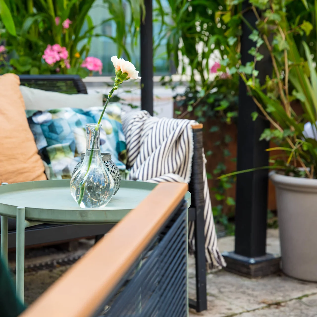 A single flower in a vase on a garden table, surrounded by plants and outdoor furniture.