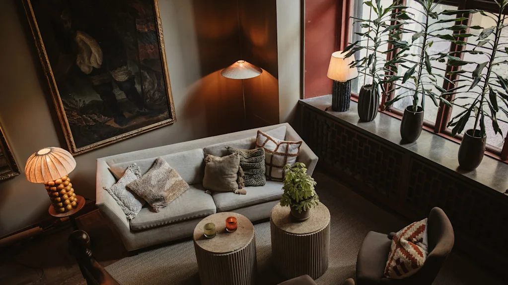 A cozy living room with a beige sofa adorned with cushions, two round tables, a floor lamp, tall potted plants by a window, and artwork on the wall.