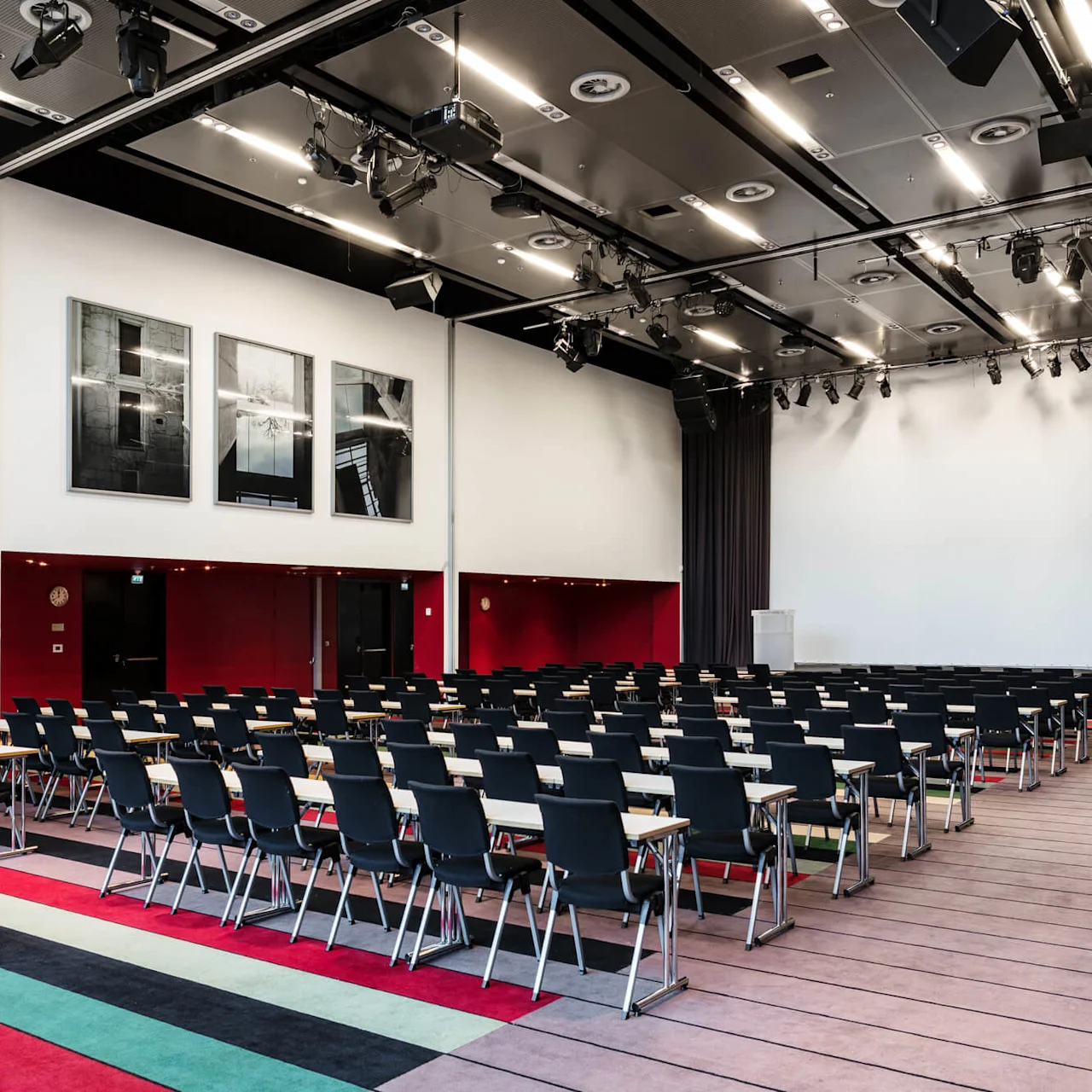 Rows of black chairs face a lectern in a large, modern conference room; walls display black and white artwork, and colorful carpet strips line the floor.