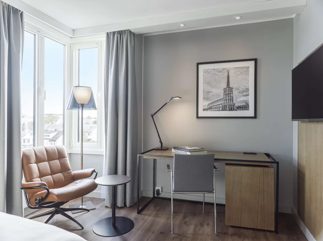 Lyst hotellrom med skrivebord, kontorstol, en behagelig skinnlenestol ved vinduet og et bilde på veggen.