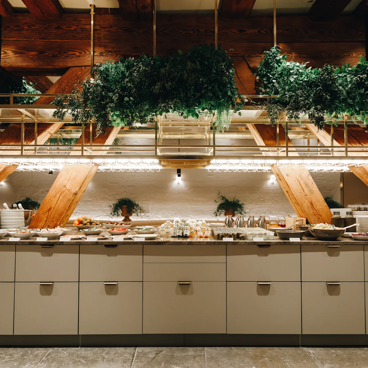 Buffet table displays various dishes with plates stacked on the side. Hanging plants and wooden beams create a rustic atmosphere.