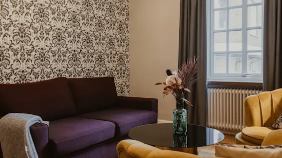 Ett mysigt hotellrum på Home Hotel Grand, Helsingborg, med en lila soffa, gul fåtölj och ett soffbord med blommor.