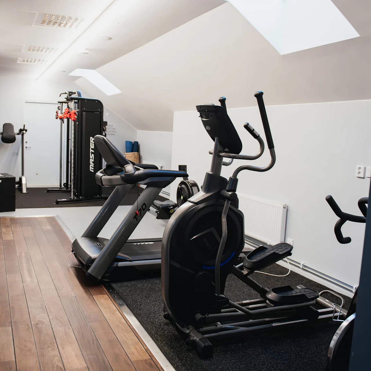 Treadmill and elliptical positioned on a wooden floor in a well-lit gym room, equipped with various exercise machines in the background. Text: "MASTER."