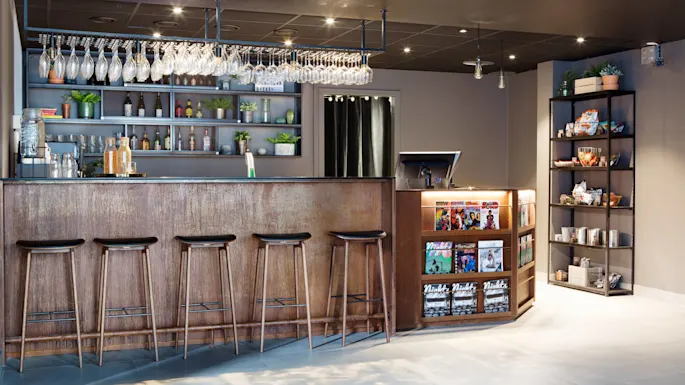 Bar with wooden stools stands in front of shelves holding glasses and bottles; nearby, a small magazine rack offers reading material in a warmly lit, modern interior.