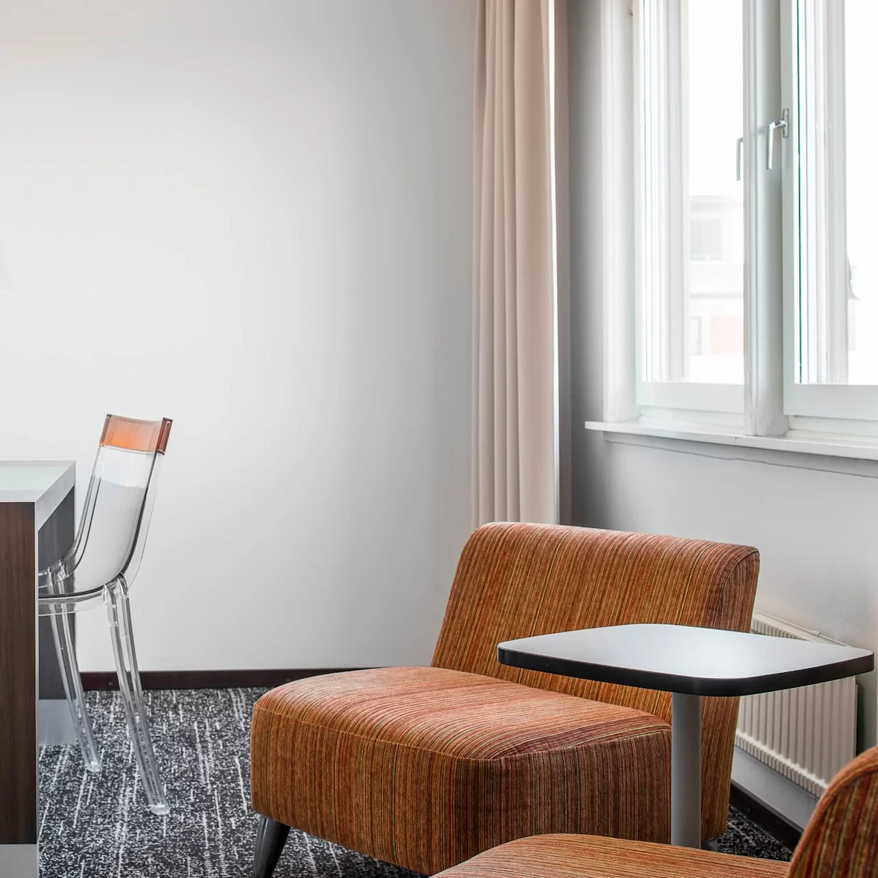 Ett mysigt rum med ett skrivbord, genomskinlig stol och lampa, bredvid orange klädda stolar och ett litet bord vid ett fönster med beige gardiner.