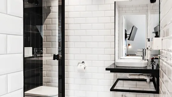 Et moderne badeværelse har en elegant vask på en sort bordplade, omgivet af hvide fliser. En glasbruser er ved siden af, med et vægmonteret TV synligt i spejlet.