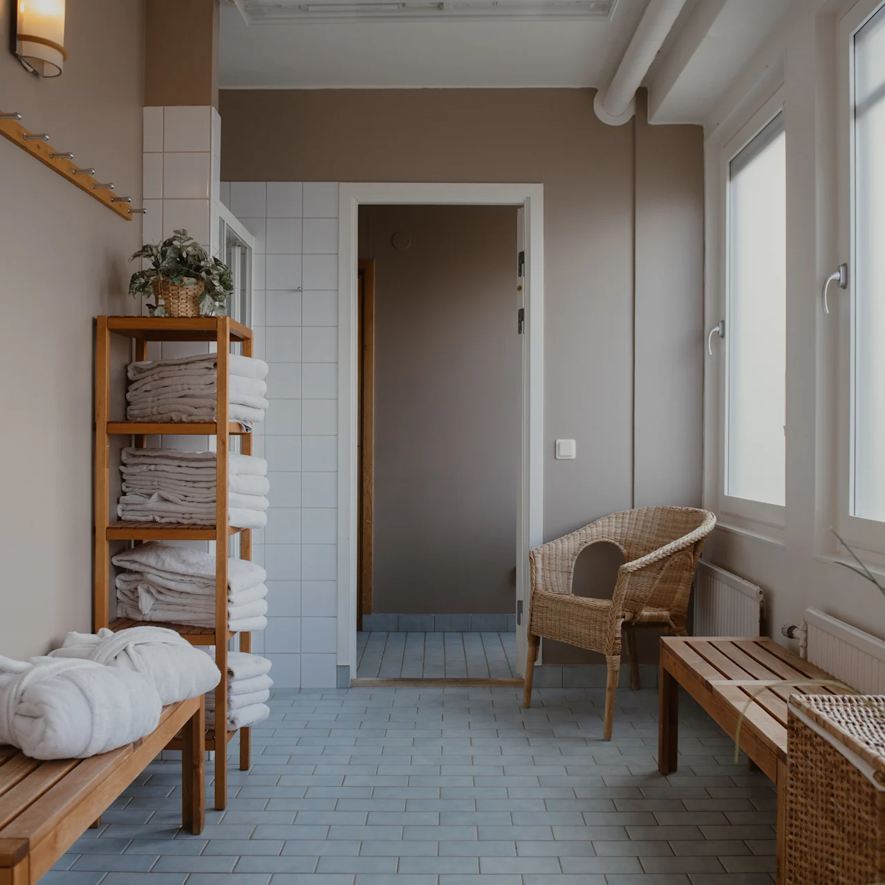Wooden benches and shelves hold folded towels in a corridor with beige walls, adjacent to a wicker chair, soft lighting, and large frosted windows.