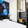 A hotel room features a double bed beneath a wall mural of a guitarist on stage. A wardrobe with shelves and a yellow suitcase is on the right.