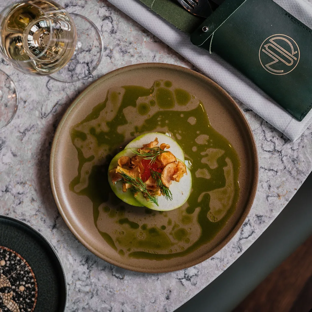 Tyylikkäästi aseteltu annos, jossa on vihreää kastiketta, valkoinen pohja, rapeita lisukkeita ja tuoreita yrttejä, viinilasien kera.