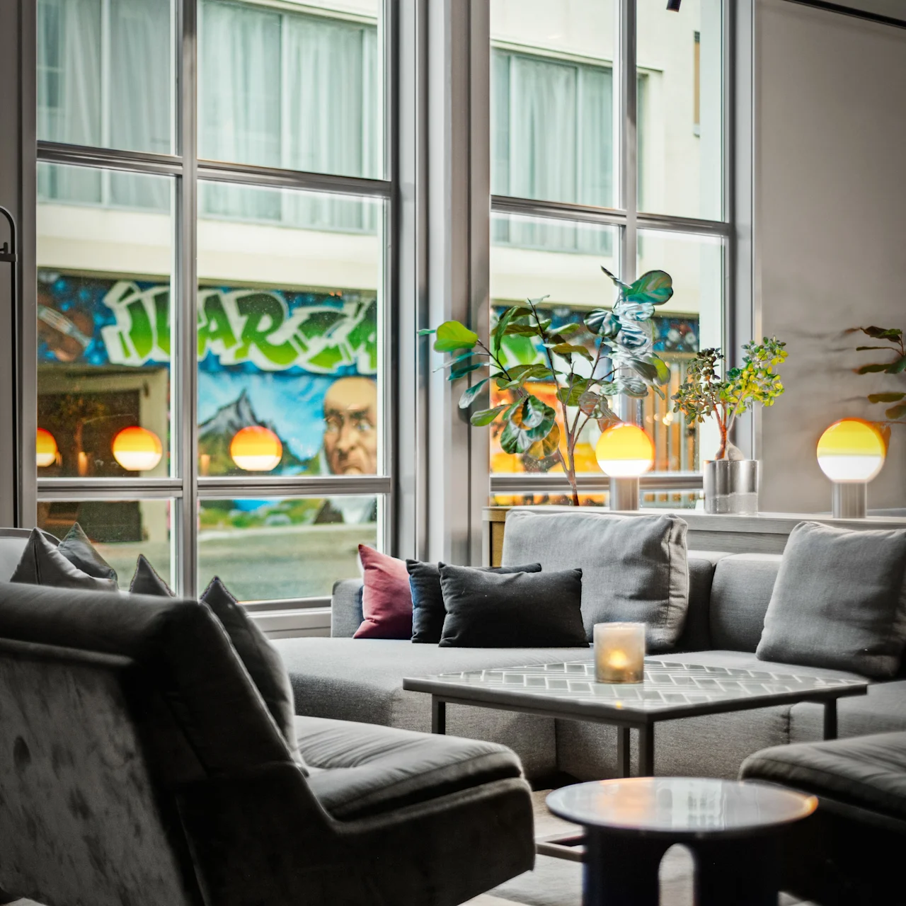 En moderne stue har grå sofaer og lænestole omkring et bord med et lys, store vinduer med urban graffiti udenfor, og frodig grønt interiør.
