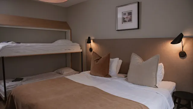 En säng med beige täcken och kuddar, belägen nära en våningssäng, i ett svagt upplyst rum med vägglampor och inramad konst ovanför sänggaveln.