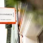 A rectangular sign reads "HAMMER HOME HOTEL" in black text, bordered by orange, hanging on a building's exterior wall against a cloudy sky.