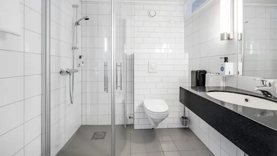 Brusekabine lukket i glasdør står mod flisevæg. Tilstødende toilet og sort bordplade med vask er oplyst af vægmonterede lys. Hvidt og minimalistisk design.