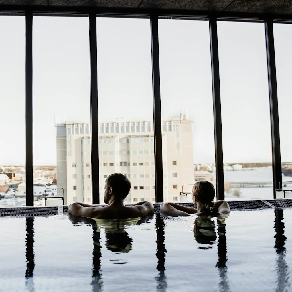 Kaksi henkilöä rentoutuu sisäuima-altaassa katsellen suurista ikkunoista kaupunginäkymää, luoden rauhallisen ja ylellisen ilmapiirin.