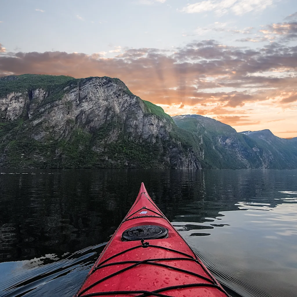 En rød kajakk glir over rolig vann, omgitt av bratte, grønne fjell, under en solnedgangshimmel med spredte skyer.