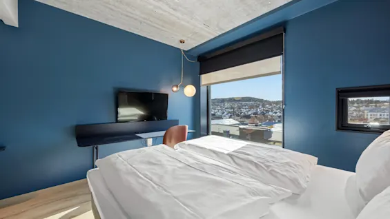 Et moderne hotellrom med en komfortabel seng, et skrivebord med TV og et stort vindu med utsikt over en by.