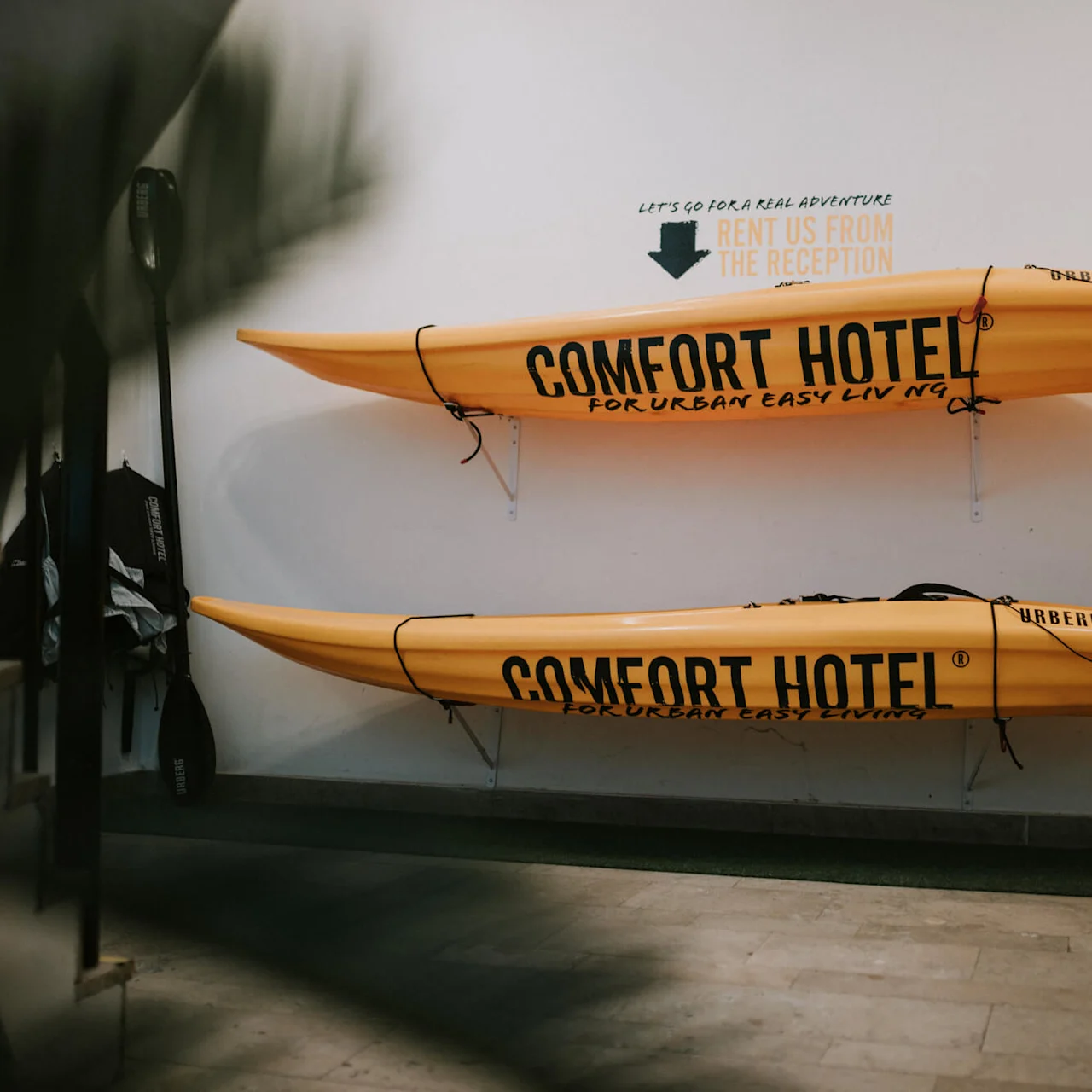 To gule kajakker monteret på en væg med pagajer, mærket "COMFORT HOTEL". Et skilt ovenfor lyder "LET'S GO FOR A REAL ADVENTURE RENT US FROM THE RECEPTION COMFORT HOTEL FOR URBAN EASY LIVING".